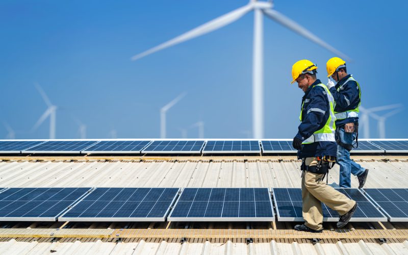 Worker Technicians are working to construct solar panels system on roof with wind turbine on background. Installing solar photovoltaic panel system. Renewable clean energy technology concept.